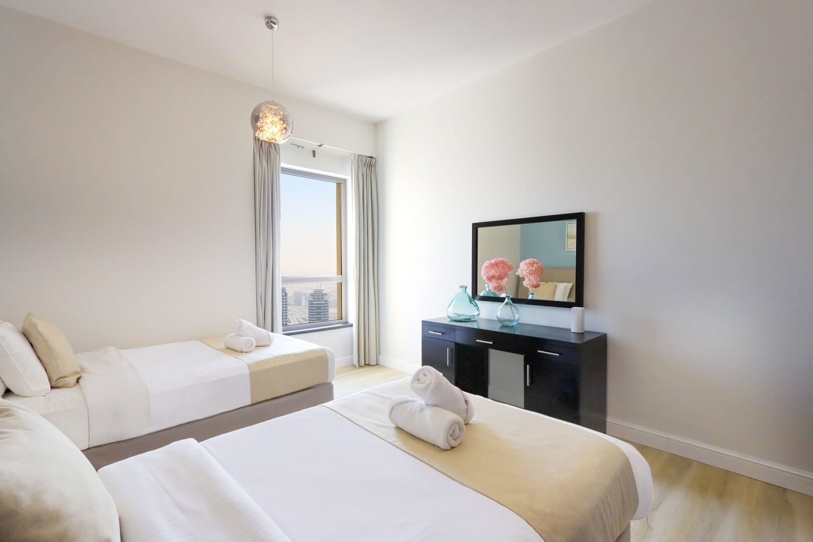 Quarto de hotel com duas camas, uma janela grande com cortinas, uma cômoda preta com espelho e um lustre.