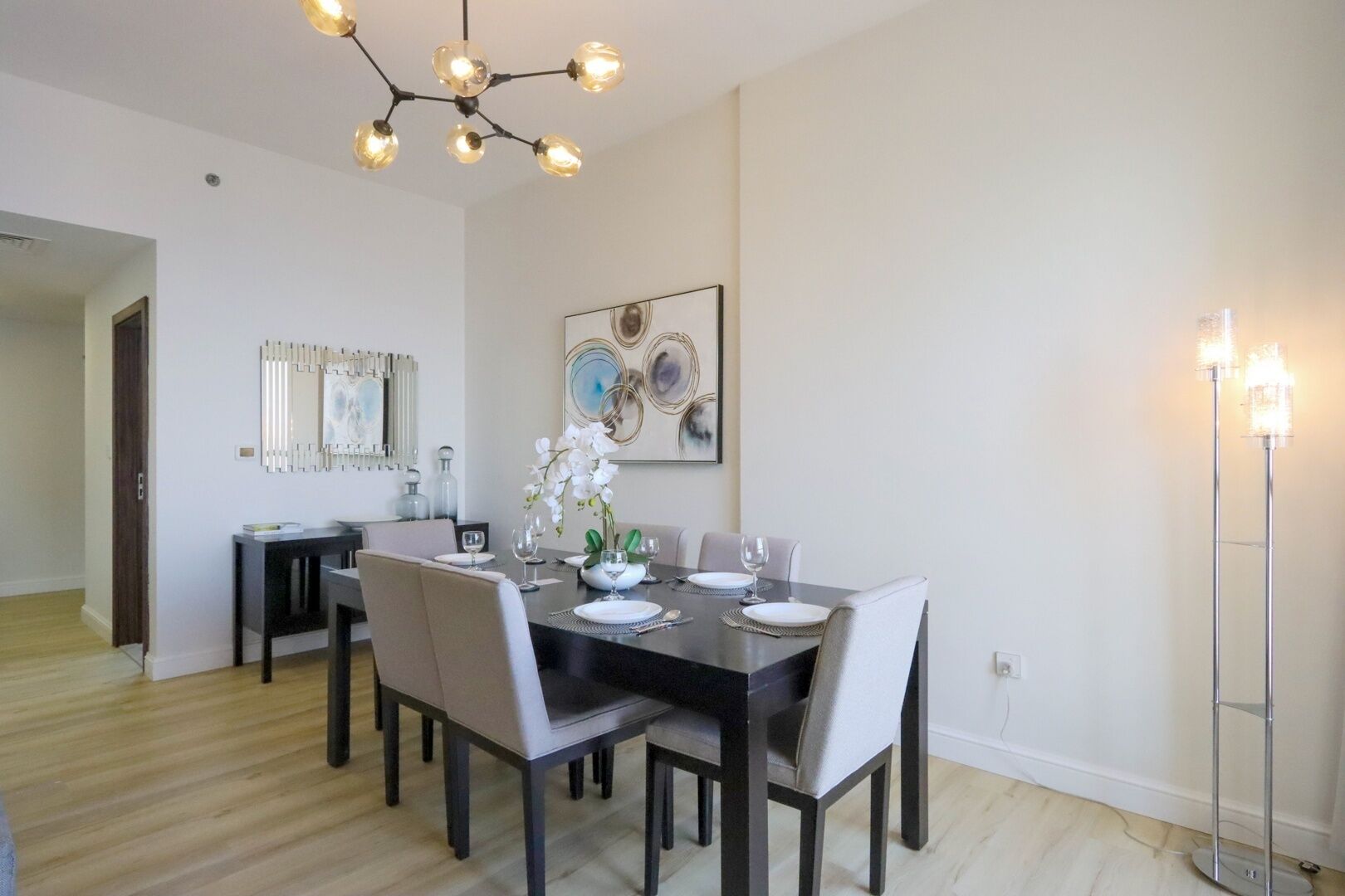 A dining area with a dark wooden table set for four, surrounded by chairs. There is a modern chandelier above the table and a floor lamp to the right.