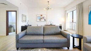 A modern living room with a grey sofa, a round side table, a chandelier, and a city view through large windows. - Livbnb High Floor 2B  Marina View | Walk to Beach (Dubai)