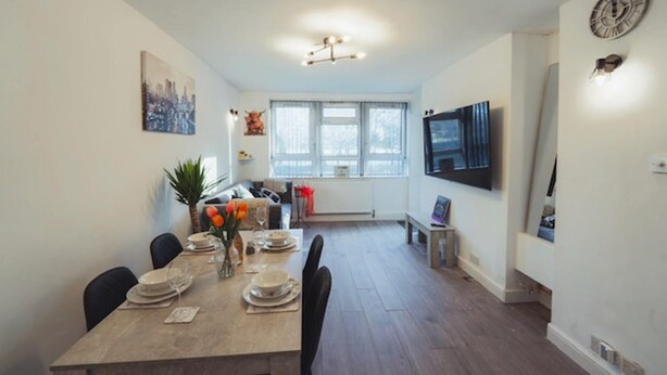 A dining area with a long table set for four, a TV mounted on the wall, and a window with a view of trees. - 1 Bed-park View-balcony-wifi-pet Friendly (London)