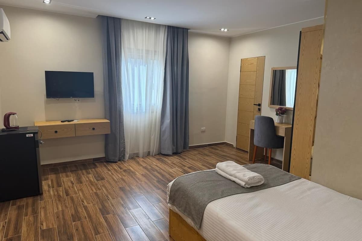 A modern hotel room with a wooden floor, a bed with a grey and white duvet, a desk with a chair, a flat-screen TV, and a large window with sheer curtains.