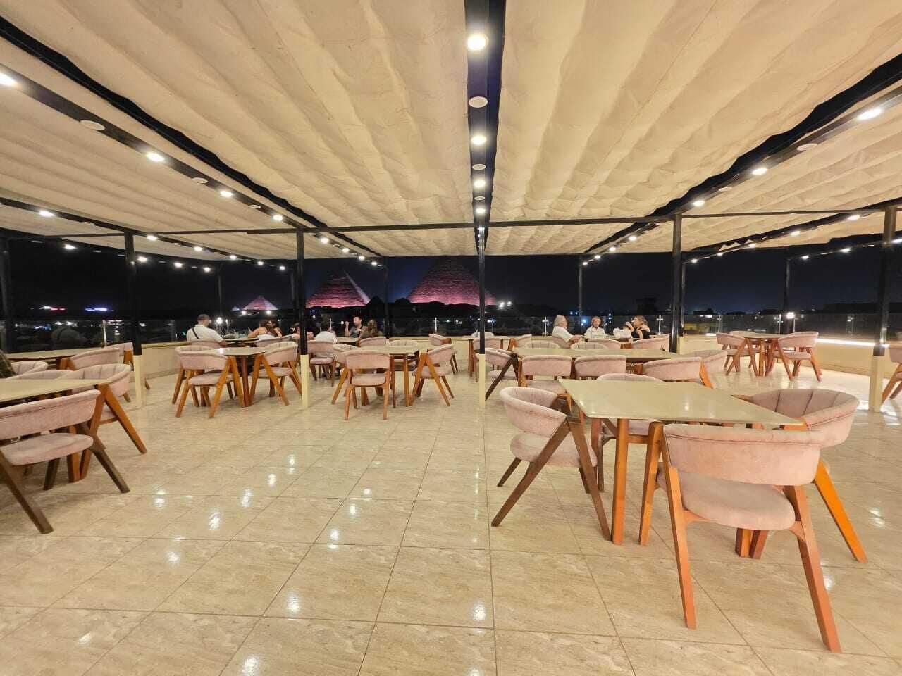 An outdoor dining area with round tables and cushioned chairs, under a covered structure with a view of the cityscape at night.