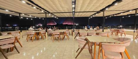 An outdoor dining area with round tables and cushioned chairs, under a covered structure with a view of the cityscape at night.