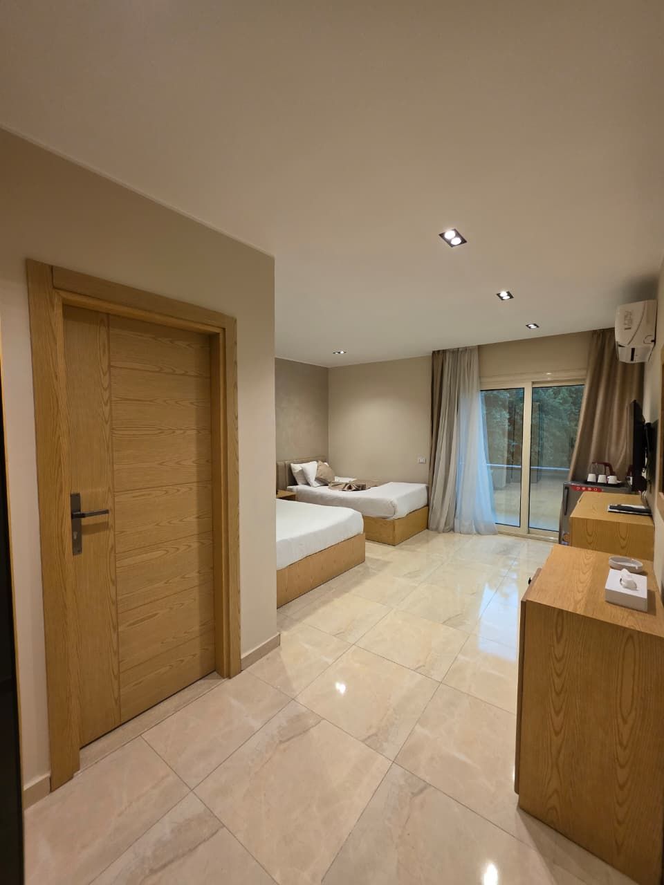 Habitación de hotel con puerta de madera, dos camas, un escritorio y un ventanal con cortinas.