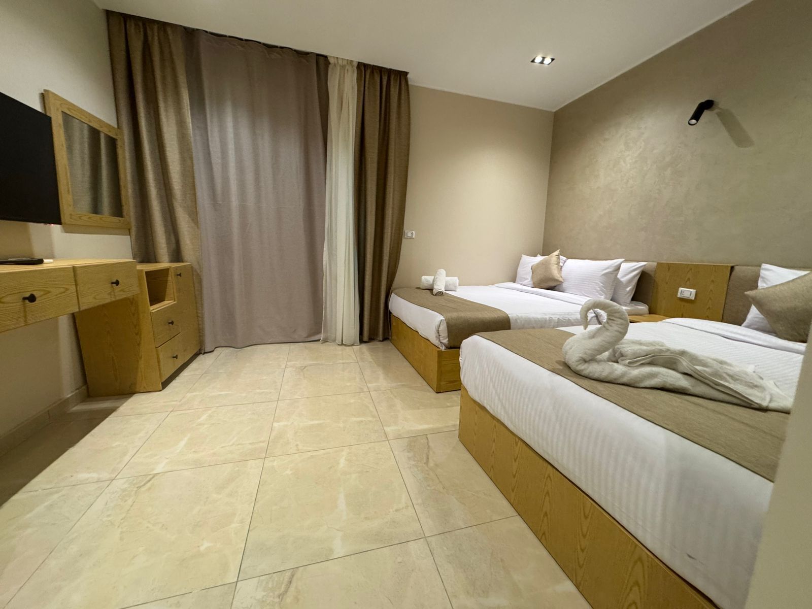 A hotel room with two beds, a wooden desk, a television, and a large window with curtains.