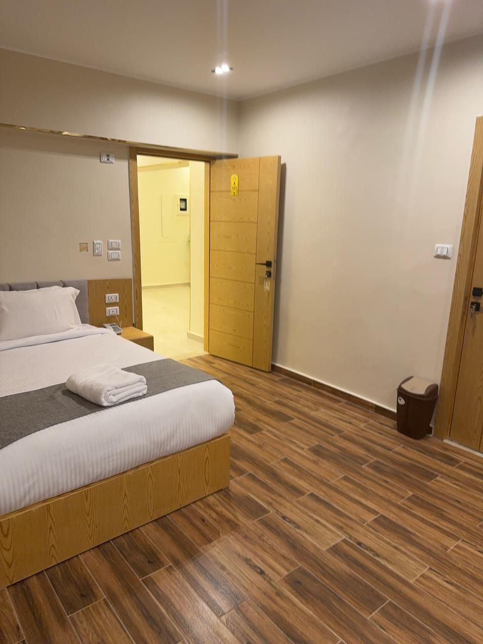 Una habitación de hotel con una cama de madera, una mesita de noche, un cubo de basura y una puerta que da a otra habitación.