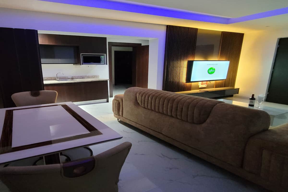 A modern living room with a brown sofa, a dining table with chairs, a flat-screen TV mounted on the wall, and a kitchen area in the background.