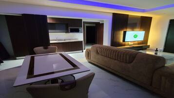 A modern living room with a brown sofa, a dining table with chairs, a flat-screen TV mounted on the wall, and a kitchen area in the background.
