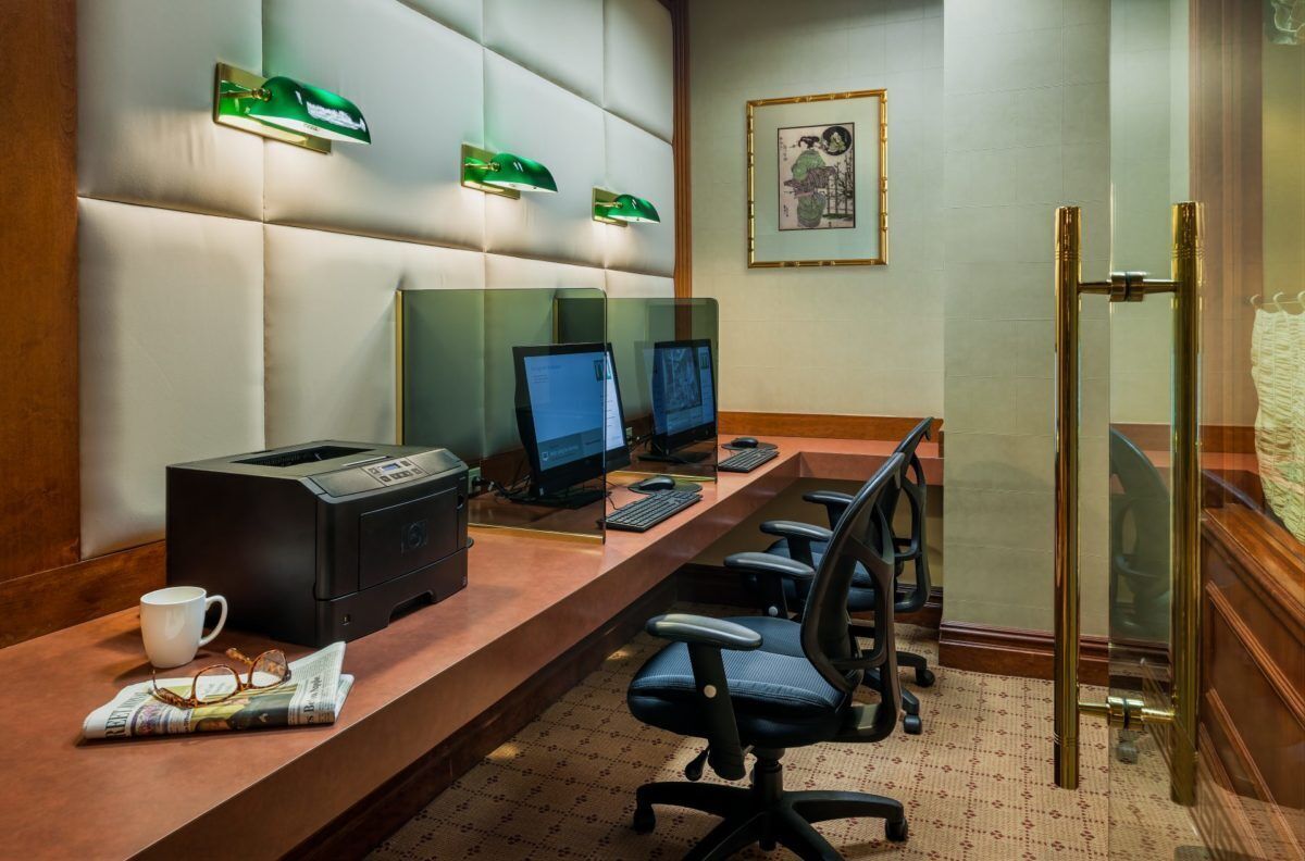 A hotel room with a long desk, two computers, a printer, and a pair of chairs.