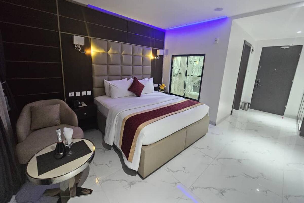 A modern hotel room with a large bed, a small round table with a bottle and glass, a chair, and a large window with a city view.