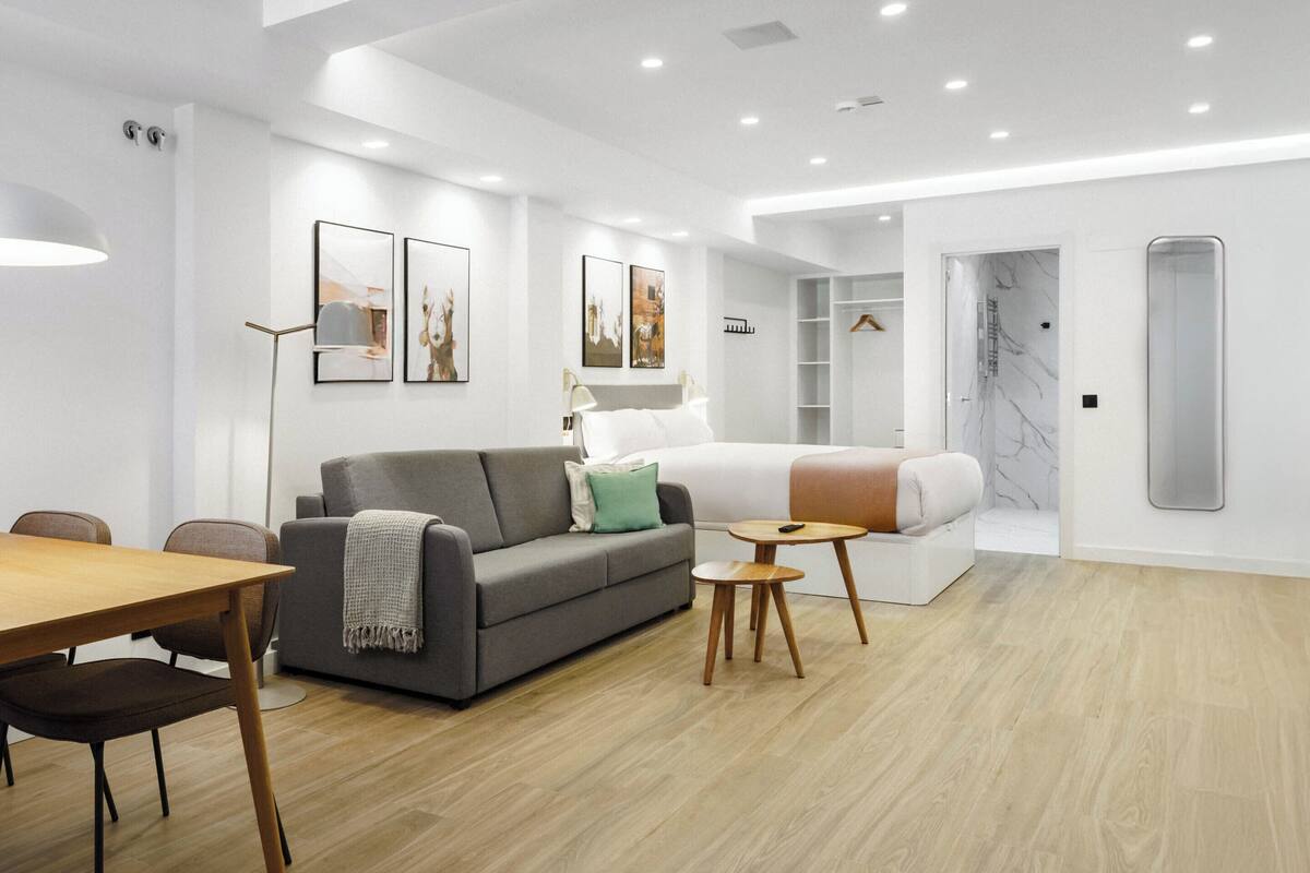 A modern living room with a grey sofa, a small round table, and a wooden dining table. There is a bed with a white headboard and a marble wall in the background.
