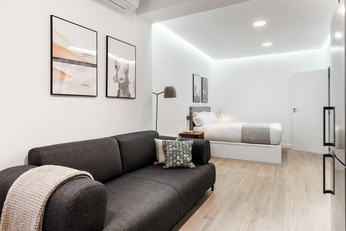 A modern, compact hotel room with a grey sofa, a bed with a grey headboard, and two framed pictures on the wall.