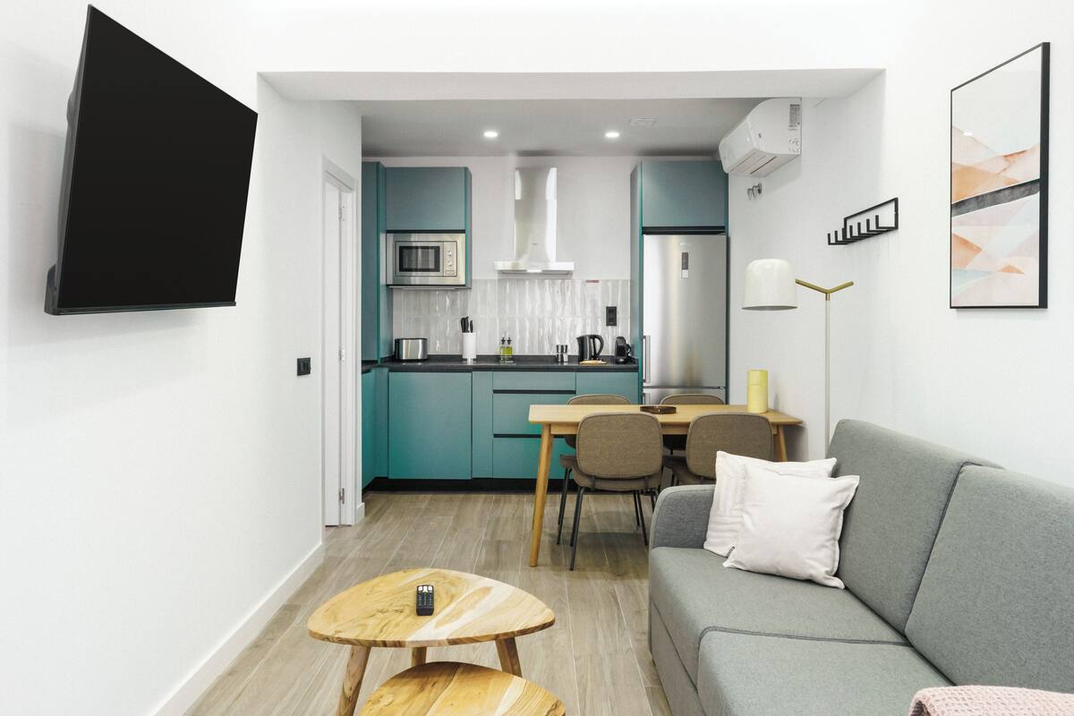 A modern kitchen with teal cabinets, a stainless steel refrigerator, and a microwave. There is a dining area with a gray sofa, a small wooden table, and a floor lamp.