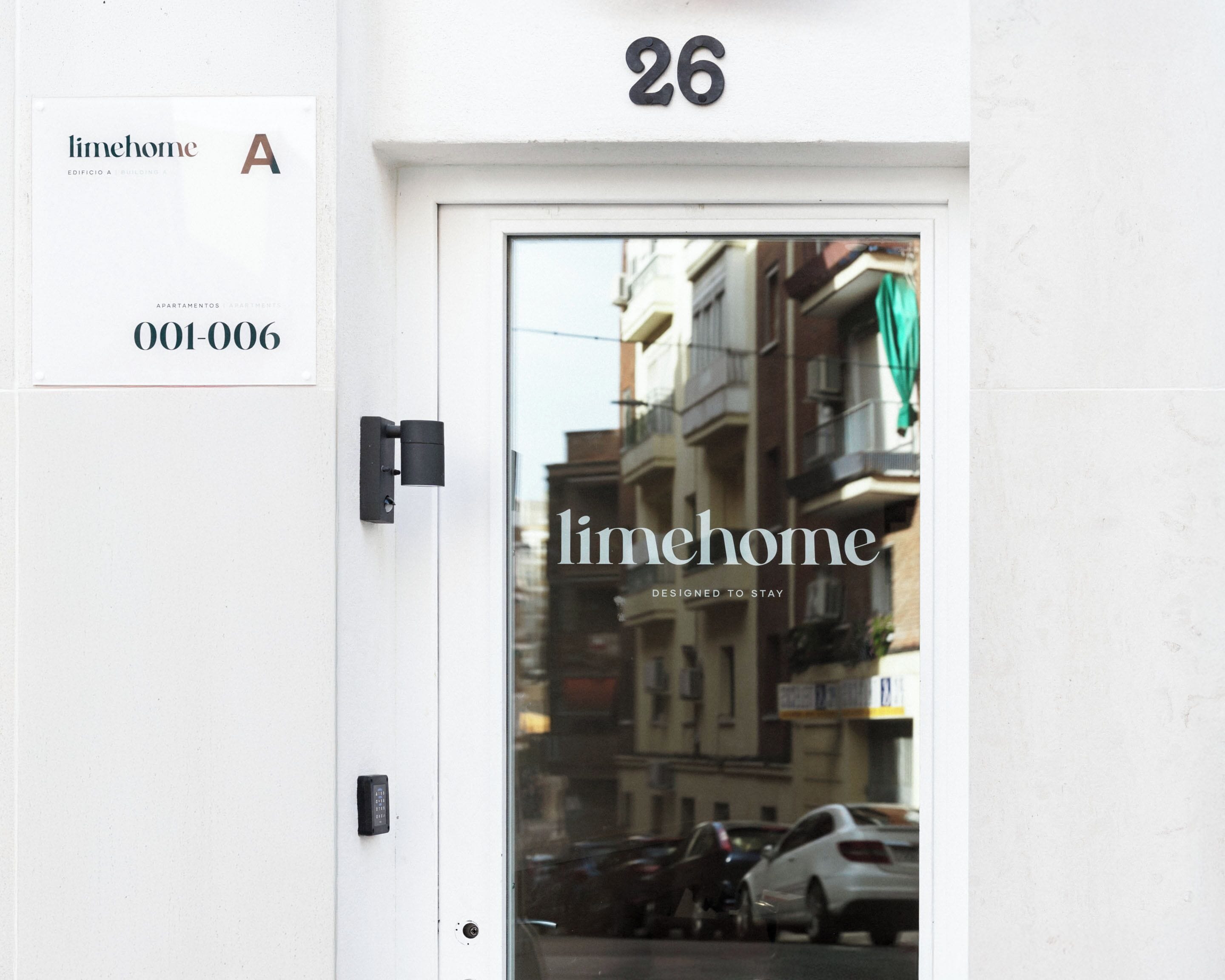 Una puerta con el número 26, un cartel que indica 'limehome' apartamentos, y una vista parcial de coches aparcados y edificios al fondo.