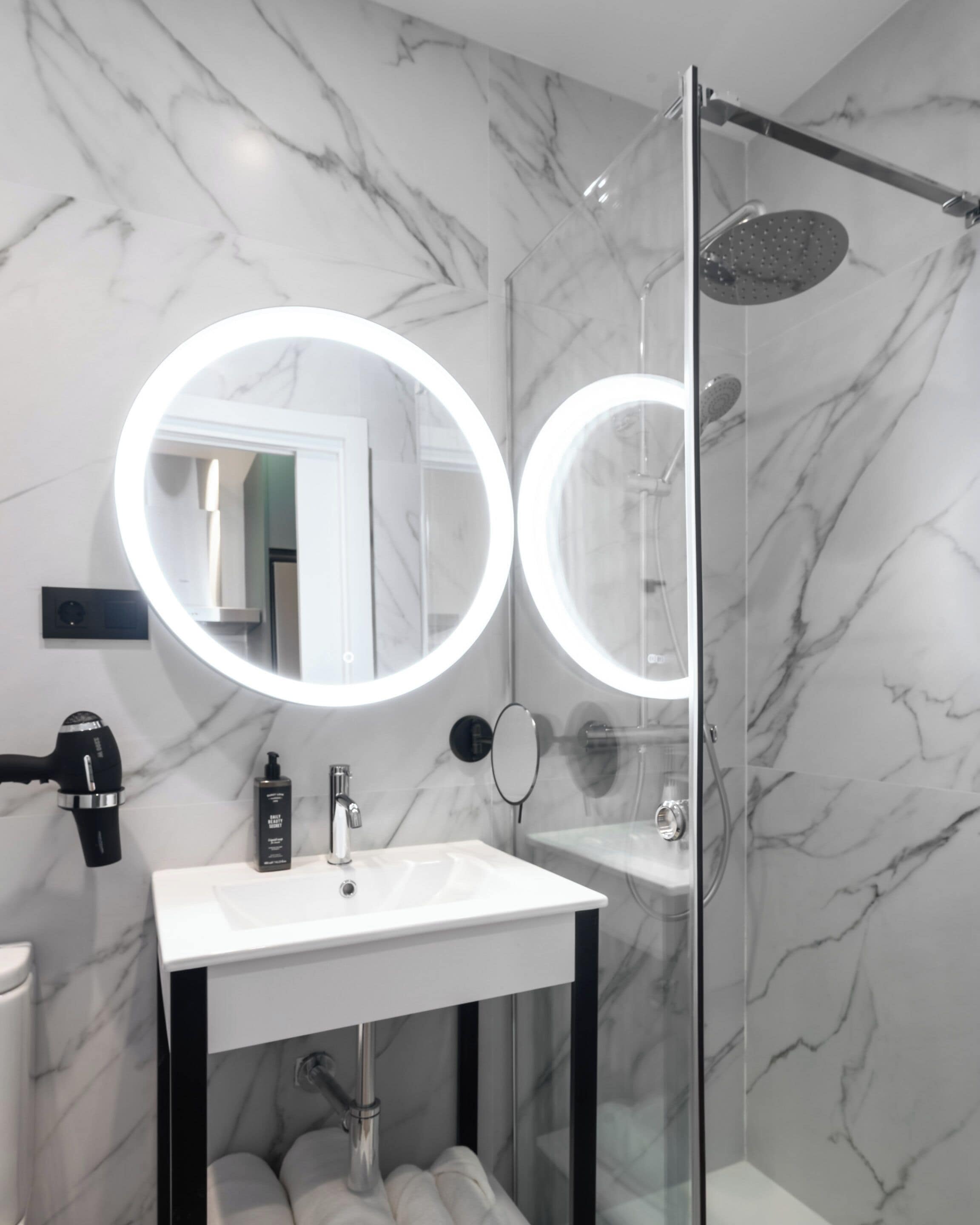 A modern bathroom with a glass-enclosed shower, a white sink, and two round mirrors with built-in lighting.