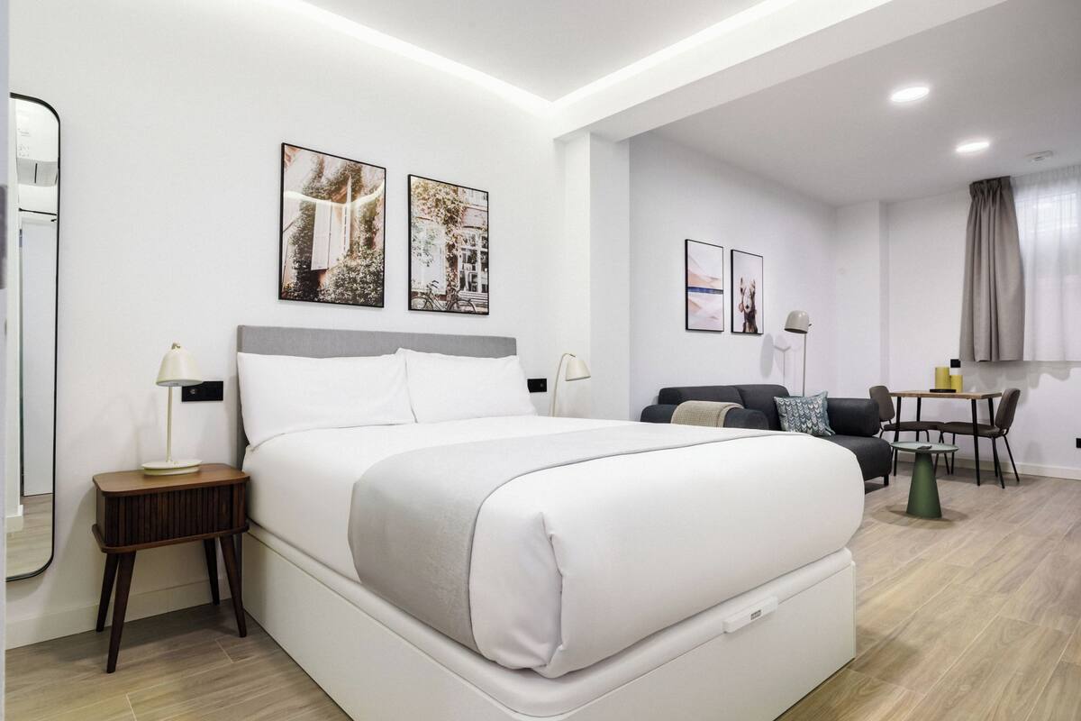 A modern hotel room with a large bed, a small side table, a sofa, a dining table, and a mirror.