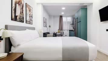 Quarto de hotel moderno com uma cama grande, mesa de cabeceira, escrivaninha e uma cadeira. Cozinha compacta ao fundo.