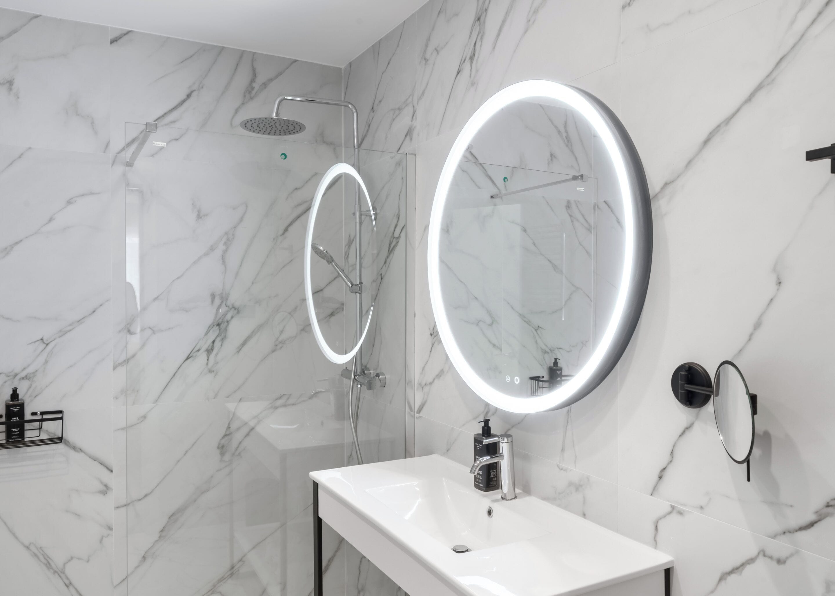 A modern bathroom with a marble wall, a shower with a handheld showerhead, a large mirror with a light, and a white sink with a soap dispenser.