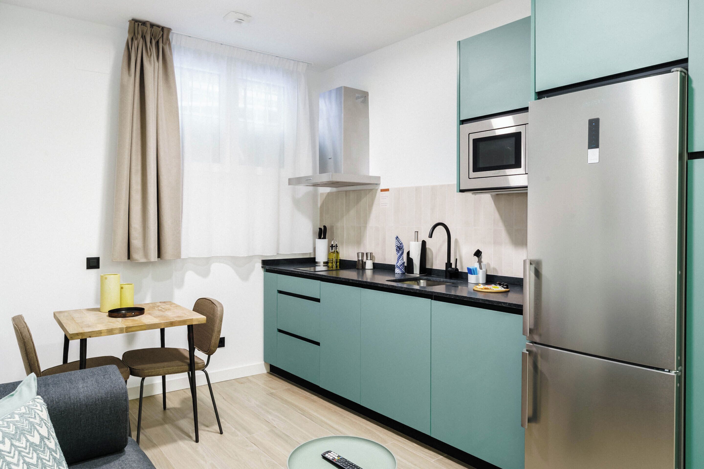 A compact kitchen with teal cabinets, a stainless steel refrigerator, a microwave, and a dining area with a wooden table and chairs.