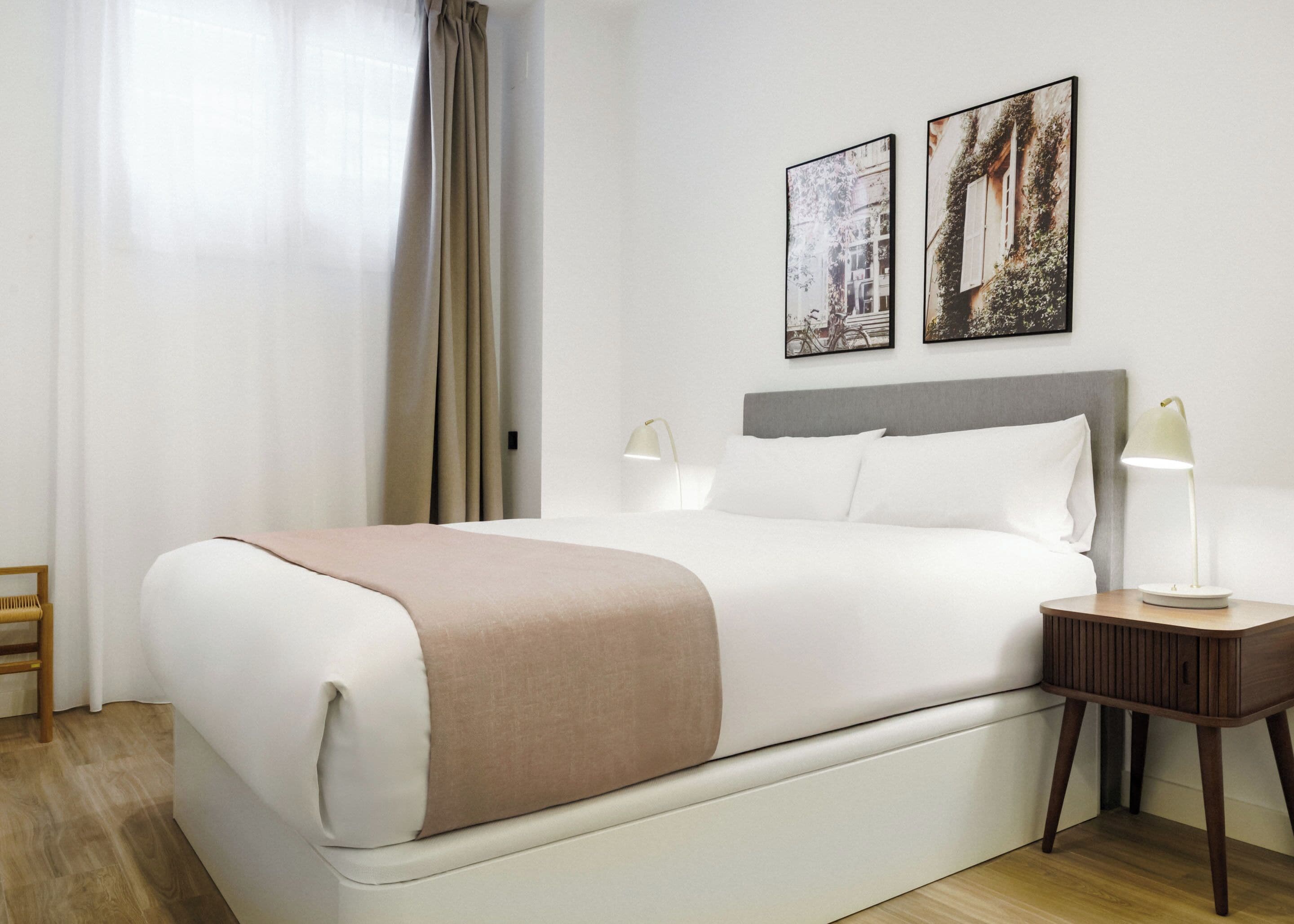 A neatly made bed with a white and beige bedspread, two wall-mounted lamps, and two framed pictures above the bed.