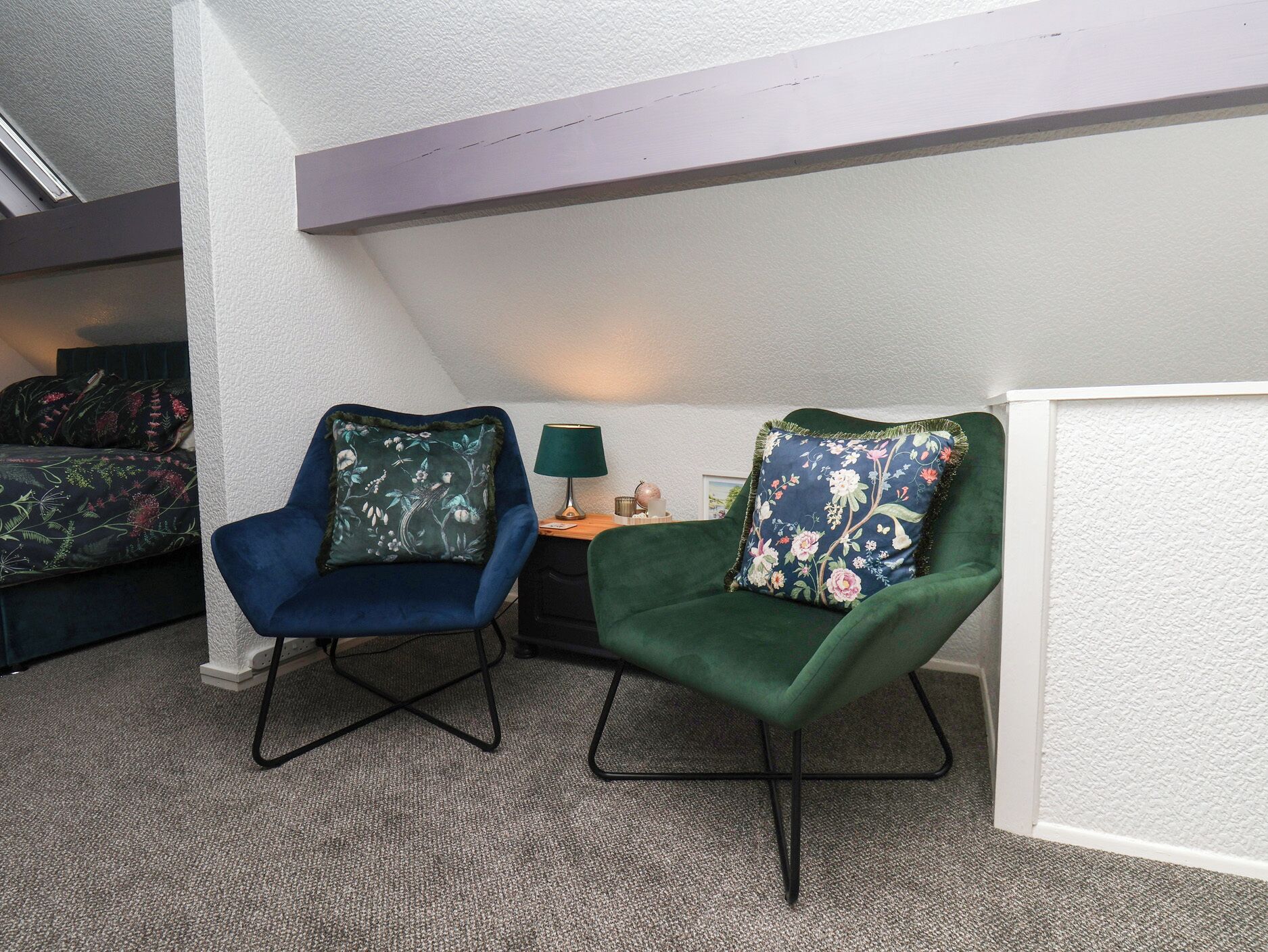 Una habitación pequeña y acogedora con dos sillones, uno azul y otro verde, cada uno con cojines estampados, una mesita con una lámpara, y una cama al fondo.