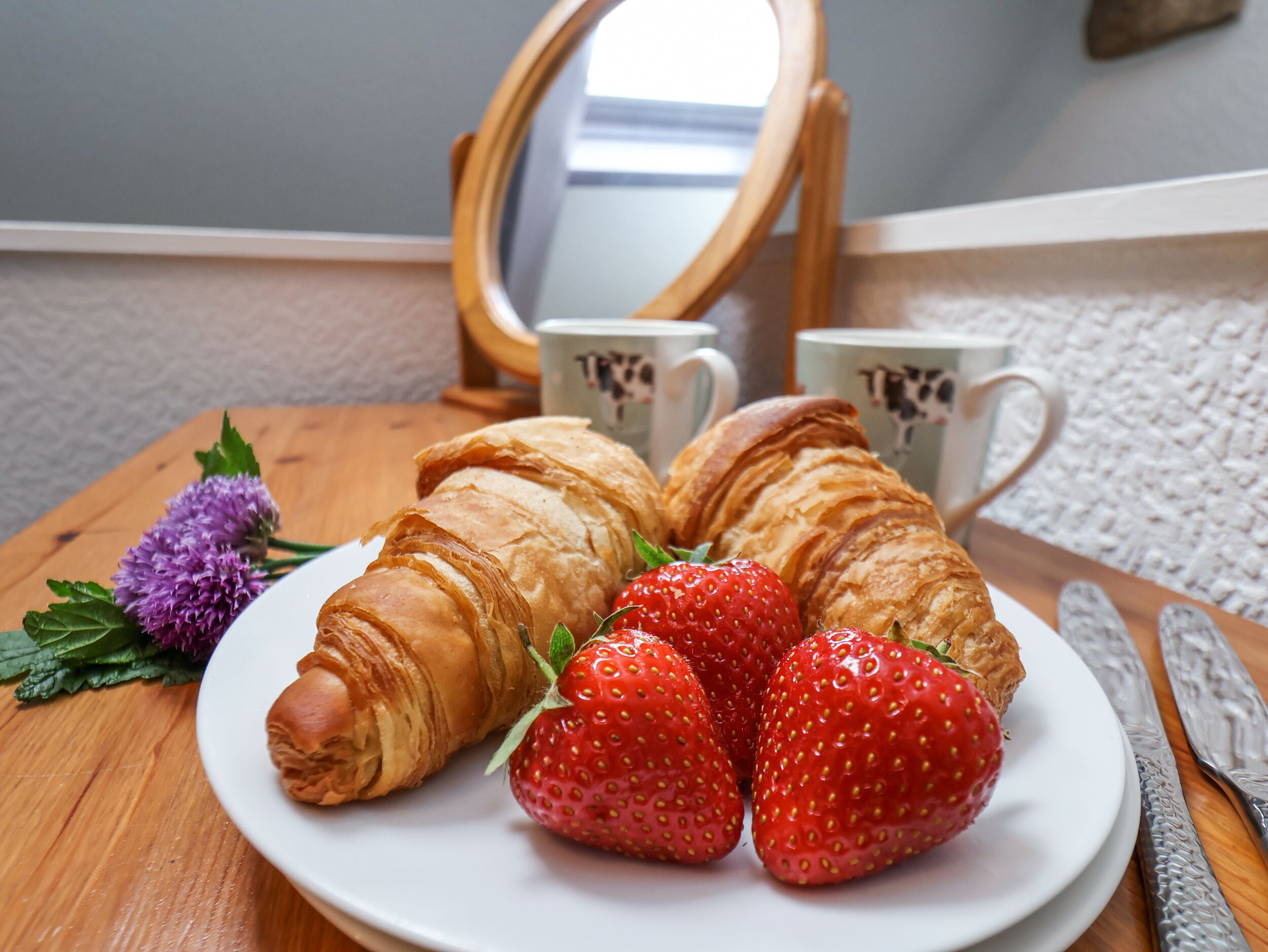A plate with croissants and strawberries on a wooden table, with a mirror and two mugs in the background.