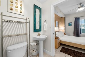 A modern bathroom with a toilet, sink, and towel rack. There is a bed with a nightstand and lamp in the adjacent room. - Cassine Station by Panhandle Getaways (Santa Rosa Beach)