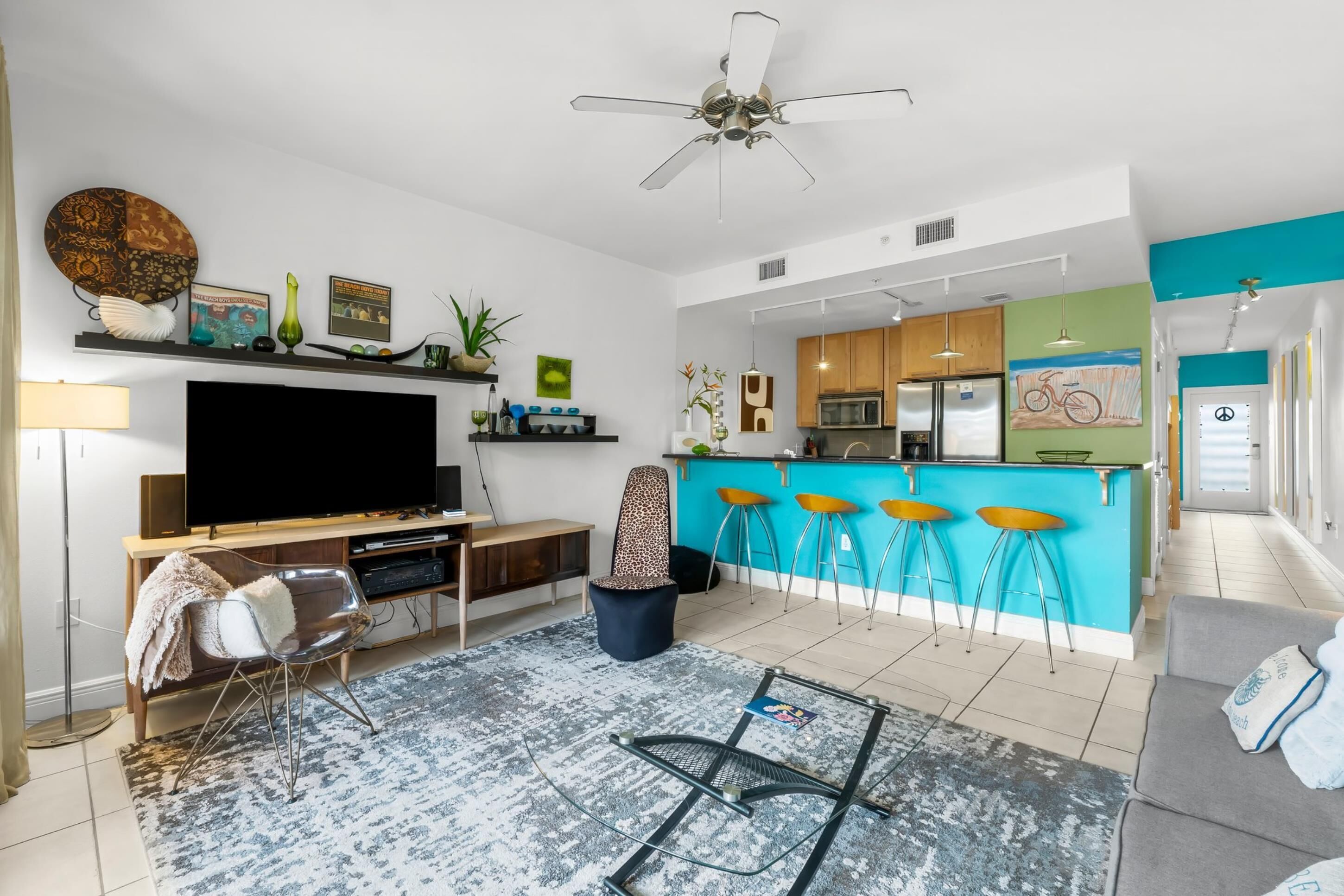 A modern living room with a flat-screen TV, a ceiling fan, and a kitchen area with a bar.