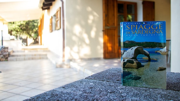 A book titled 'SPIAGGE DI SARDEGNA' on a stone ledge outside a building with a tiled floor and wooden doors. - Sardinia Re - Villa Green Val (Alghero)
