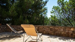 Two beige lounge chairs on a tiled patio with a stone wall and trees in the background. - Sardinia Re - Villa Green Val (Alghero)
