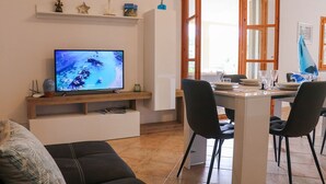 A living room with a TV, a dining table with chairs, a sofa, and a window with wooden frames. - Sardinia Re - Villa Green Val (Alghero)