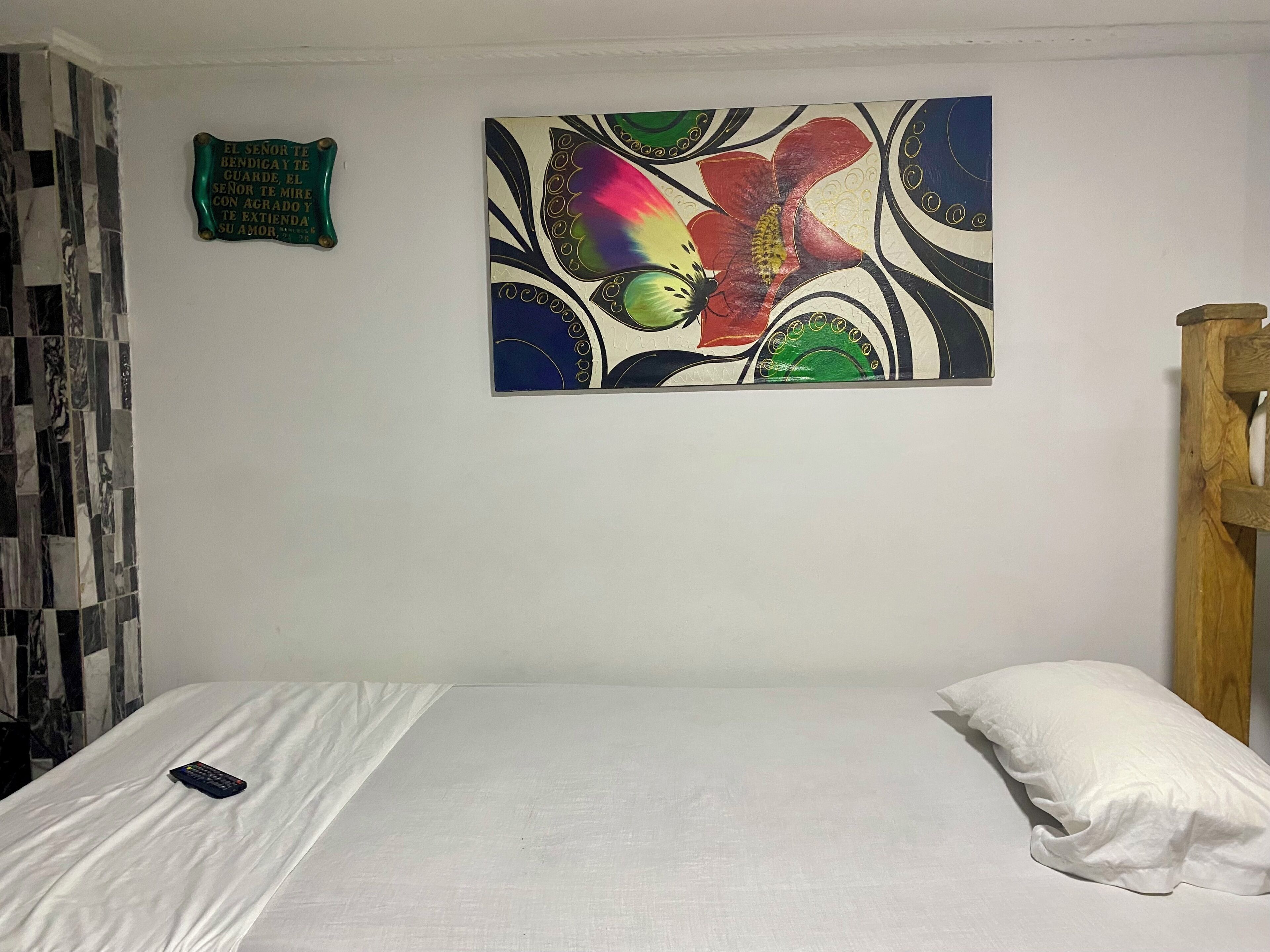 A bedroom with a bed, a pillow, a remote control, a decorative wall piece, and a small green sign.