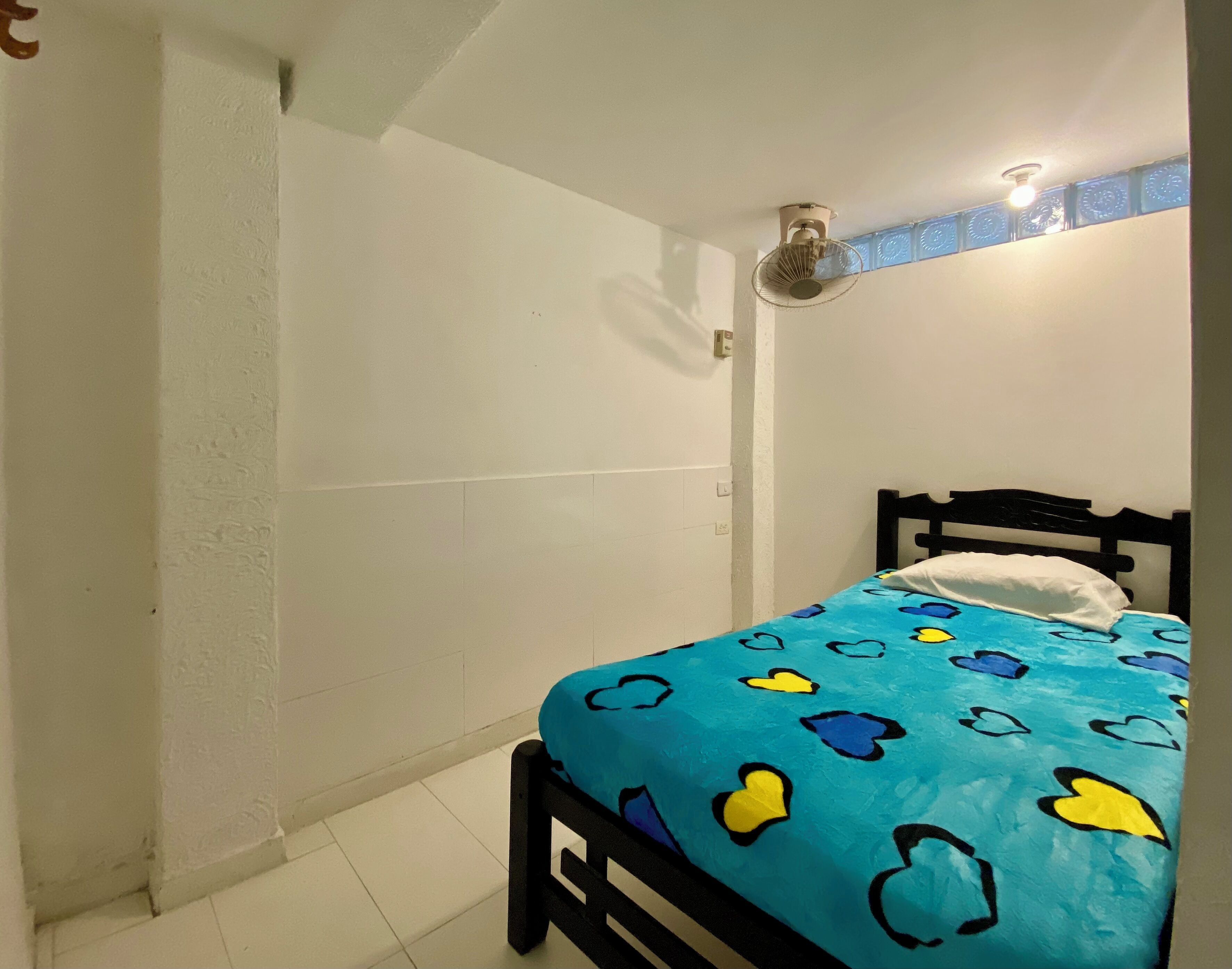 Un dormitorio con una cama, un ventilador de pared y una ventana con detalles azules decorativos.