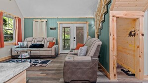 A cozy living room with a sofa, armchair, and coffee table. There is a door leading to another room and a window with curtains. - Northwoods Ln Cabin in Clear Creek by Tranquility Retreats (Sigel)