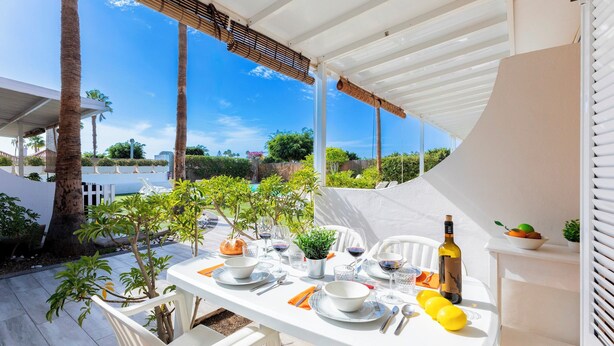 Un área de comedor al aire libre con una mesa blanca dispuesta para dos personas, rodeada de sillas blancas, una pequeña planta en maceta y una botella de vino.