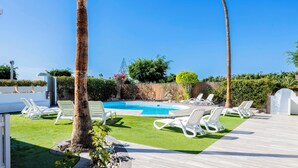 Un área de piscina con sillas de descanso blancas, cielo azul despejado y césped cuidado.