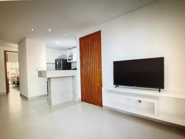 Una sala de estar moderna con un televisor de pantalla plana montado en la pared, una puerta de madera y una cocina al fondo.