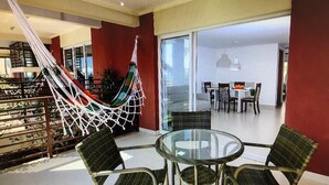 Un ambiente interno moderno con un tavolo in vetro e sedie in vimini, un’amaca e vista su una sala da pranzo attraverso ampie porte in vetro.