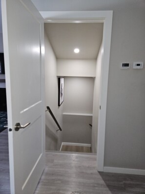 Un pasillo estrecho con una puerta a la izquierda, una escalera que sube a un nivel superior y un pequeño cuadro en la pared.