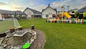 Ein Spielplatz mit Rutsche und Klettergerüsten, umgeben von einer Rasenfläche und einem Gebäude mit geneigtem Dach.