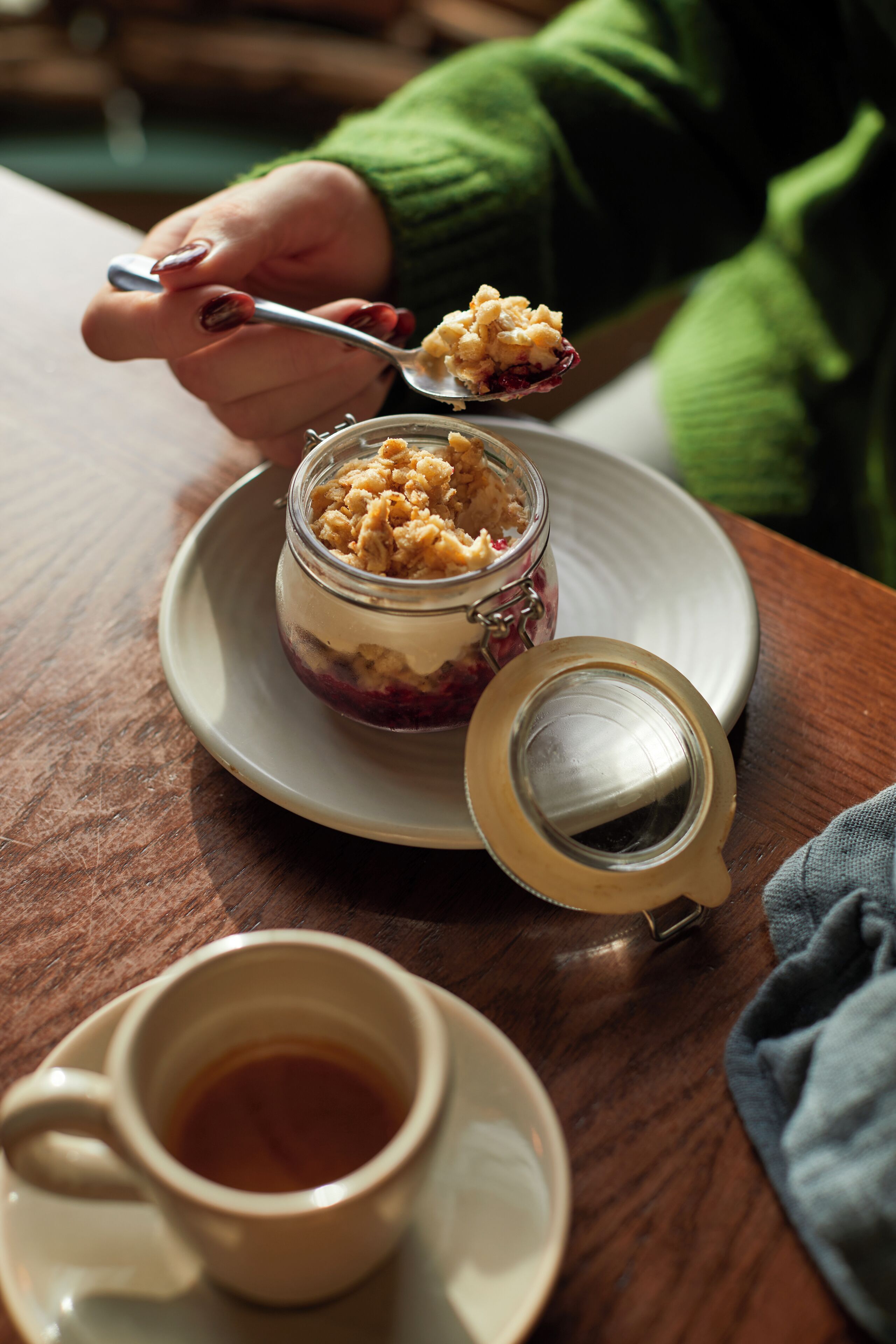 A dessert in a jar on a plate with a spoonful being lifted, a cup of tea, and a tea infuser on a wooden table.