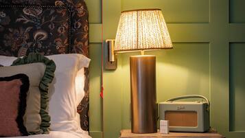 Un lit bien fait, agrémenté d’oreillers à motifs, une lampe de chevet et une radio ancienne posée sur une table de chevet en bois.