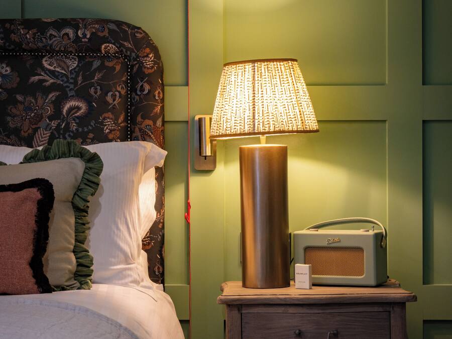Un lit bien fait, agrémenté d’oreillers à motifs, une lampe de chevet et une radio ancienne posée sur une table de chevet en bois.