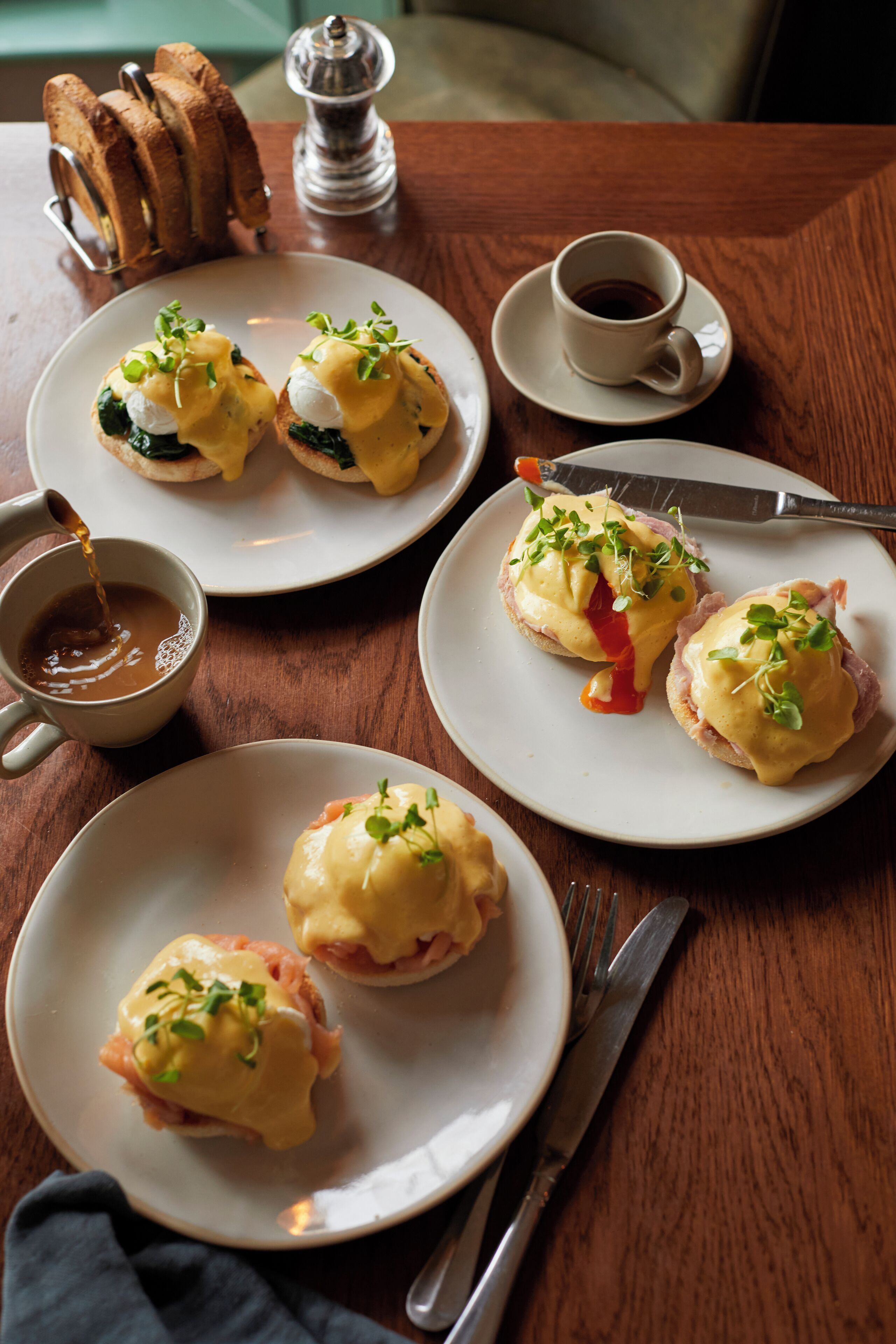 Una mesa con alimentos para el desayuno, incluyendo huevos Benedict, tostadas y café.