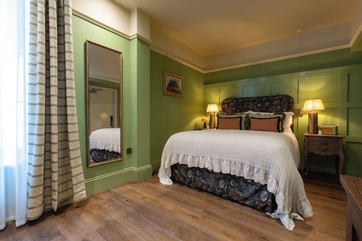 Un dormitorio con una cama grande, paredes verdes, suelo de madera, un espejo y dos lámparas de noche.