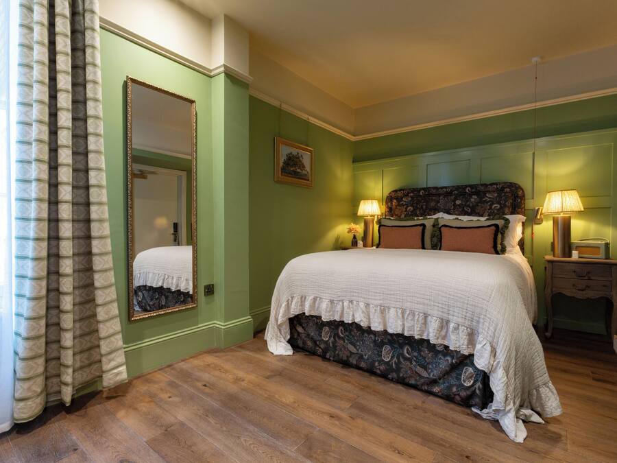 Un dormitorio con una cama grande, paredes verdes, suelo de madera, un espejo y dos lámparas de noche.