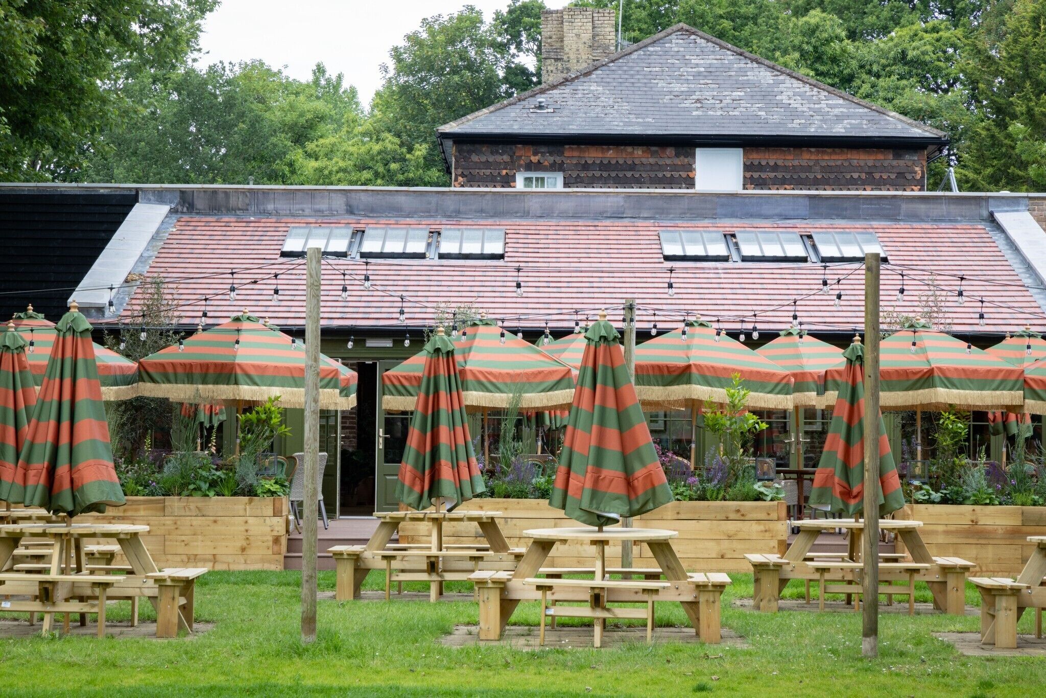 A rustic outdoor seating area with wooden tables and striped umbrellas, set against a backdrop of a traditional building with a tiled roof.