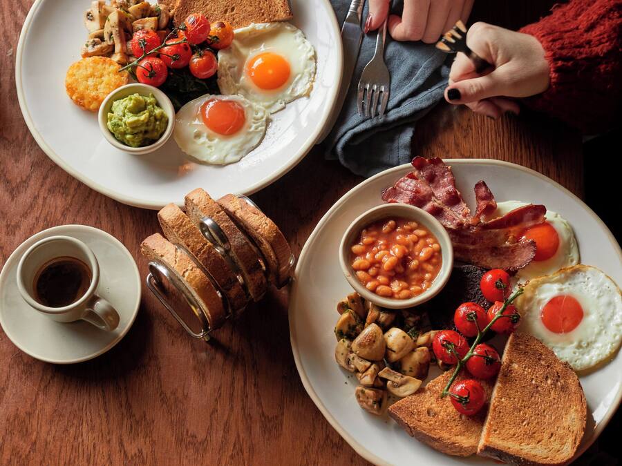 A breakfast setting with plates of eggs, bacon, beans, toast, and a cup of coffee.