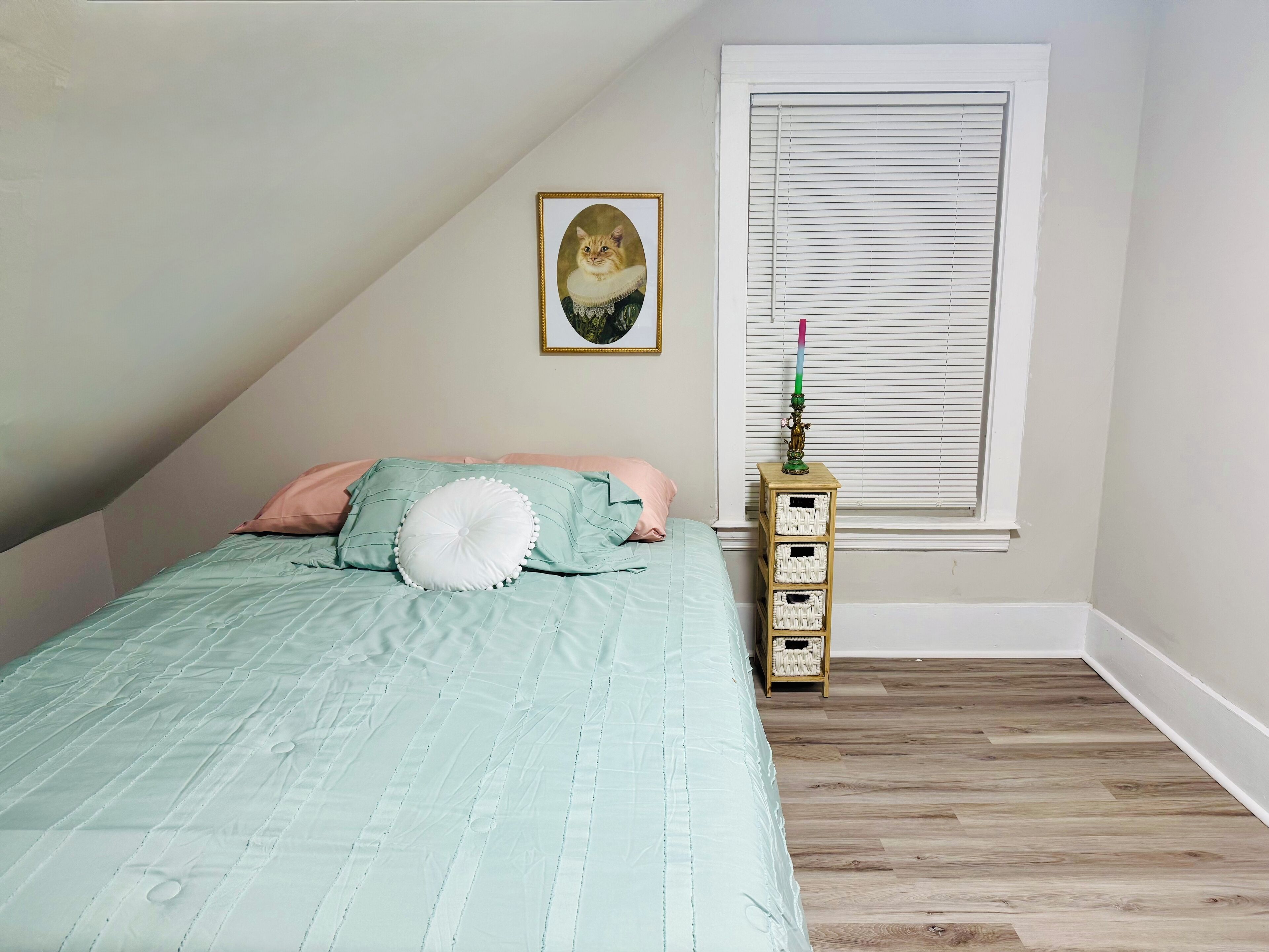 Une chambre à coucher avec un lit, des oreillers, un tableau encadré au mur, une fenêtre avec des stores et une petite armoire en bois.