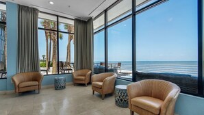 Habitación con amplias ventanas que ofrecen vistas a la playa y palmeras. Se observan varias sillas de mimbre y una pequeña mesa redonda.